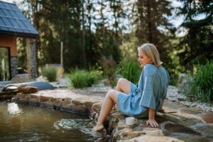 Happy young woman sitting by pond near cottege and enjoying cup of morning coffee on summer vacation